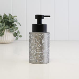 May include: A cylindrical gray soap dispenser featuring a white continuous line drawing pattern of abstract faces. The dispenser has a matte finish and is topped with a black plastic pump mechanism that extends from the center of the lid.