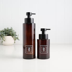 May include: Two amber glass soap dispensers with black pump tops. The larger dispenser has a rectangular label that reads "DISH SOAP" in white text. The smaller dispenser has a label that reads "HAND SOAP".