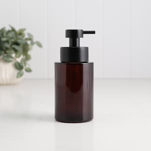 Può includere: Un dispenser di sapone in vetro marrone con una pompa nera. La bottiglia cilindrica ha una tonalità ambrata scura. La pompa è nera opaca. Il dispenser è su una superficie bianca.