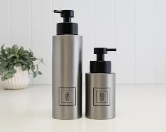 May include: Two cylindrical matte gray soap dispensers with black pump tops. The taller bottle features a square label with the text DISH SOAP, while the shorter bottle features a square label with the text HAND SOAP. Both bottles have a smooth finish.