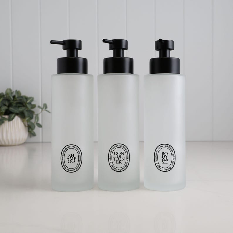 Set of three frosted glass refillable pump bottles with matte black dispensers, labelled shampoo, conditioner, and body wash in minimalist circular design. Elegant bathroom storage solution with soft matte finish, durable glass construction, and coordinated modern styling for a clean, organised shower or vanity space.