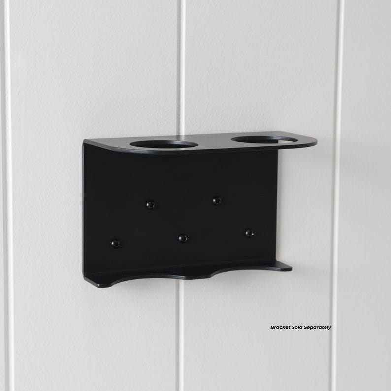 May include: A black metal wall-mounted holder with two circular openings and a small shelf. The holder has several screw holes for mounting. The text "Bracket Sold Separately" is visible at the bottom right.