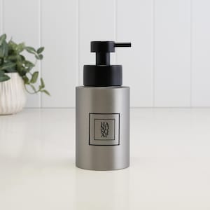 May include: A matte gray cylindrical soap dispenser with a black pump mechanism. The front features a square black border containing the stacked black text "HAND SOAP" in a clean, sans-serif font. The container has a smooth, solid finish.