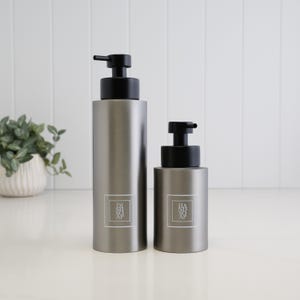May include: Two cylindrical matte grey soap dispensers with black pump heads. The taller bottle features a white square label with the text DISH SOAP, while the shorter bottle has a matching label with the text HAND SOAP printed in a clean font.