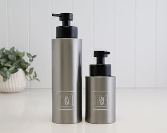 May include: Two cylindrical matte grey soap dispensers with black pump heads. The taller bottle features a white square label with the text DISH SOAP, while the shorter bottle has a matching label with the text HAND SOAP printed in a clean font.