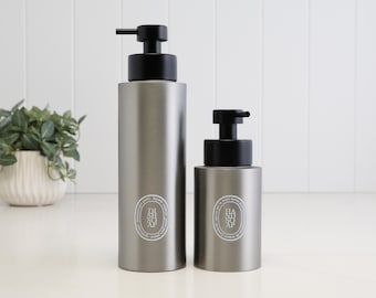 May include: Two cylindrical matte grey soap dispensers with black pump tops. The taller bottle features a white oval label reading DISH SOAP, while the shorter bottle has a white oval label reading HAND SOAP. Both containers have a smooth finish.