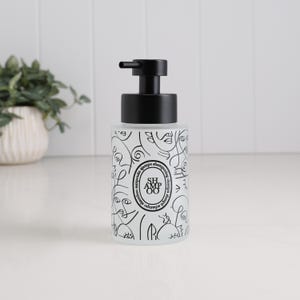 May include: A white and black shampoo dispenser with a black pump. The dispenser has a repeating line art design of faces and swirls. The word "shampoo" is printed on the bottle. The dispenser is on a white surface.