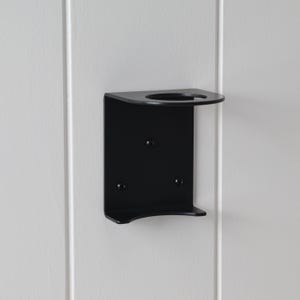 Pode incluir: Um suporte de parede de metal preto para dispensador de sabonete ou loção. O suporte tem uma abertura circular na parte superior e uma base curva. É montado em uma parede branca com painéis verticais.