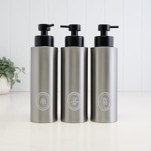 May include: Three cylindrical matte grey plastic bottles with black pump dispensers. Each bottle features a white oval label with text reading SHAMPOO, CONDITIONER, and BODY WASH respectively. The bottles are arranged in a row on a flat surface.
