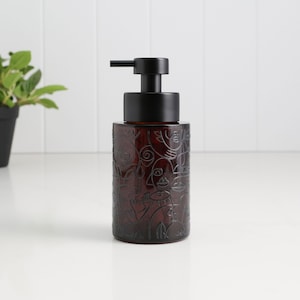 Pode incluir: Um dispensador de sabonete de vidro castanho escuro com uma bomba preta. O dispensador cilíndrico apresenta um design gravado de rostos e figuras abstratas. A bomba preta tem um bico horizontal.