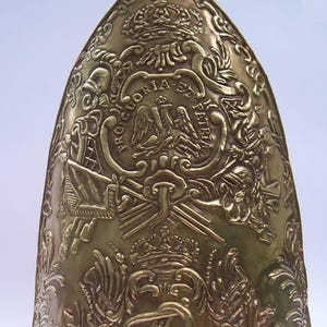 May include: A gold-colored, ornate helmet with intricate embossed designs. The helmet features a crown, eagle, and other military-themed emblems. A white and orange pompom sits atop the helmet. The helmet appears to be a historical or ceremonial piece.