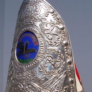 Puede incluir: Un casco plateado con intrincados diseños en relieve, incluyendo un escudo con un emblema de ave y las palabras "ET PATRIA". El casco tiene un penacho rojo y esponjoso en la parte superior.