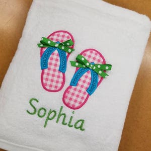 May include: White beach towel with embroidered pink and white gingham flip flops, green bows, and blue straps. The name "Sophia" is embroidered in green below the flip flops.