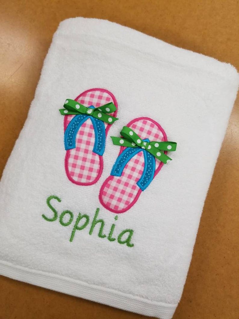 May include: White beach towel with embroidered pink and white gingham flip flops, green bows, and blue straps. The name "Sophia" is embroidered in green below the flip flops.