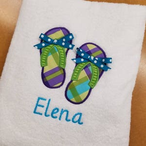 May include: White bath towel with embroidered green and purple flip flops with blue bows and white polka dots. The name "Elena" is embroidered below the flip flops.