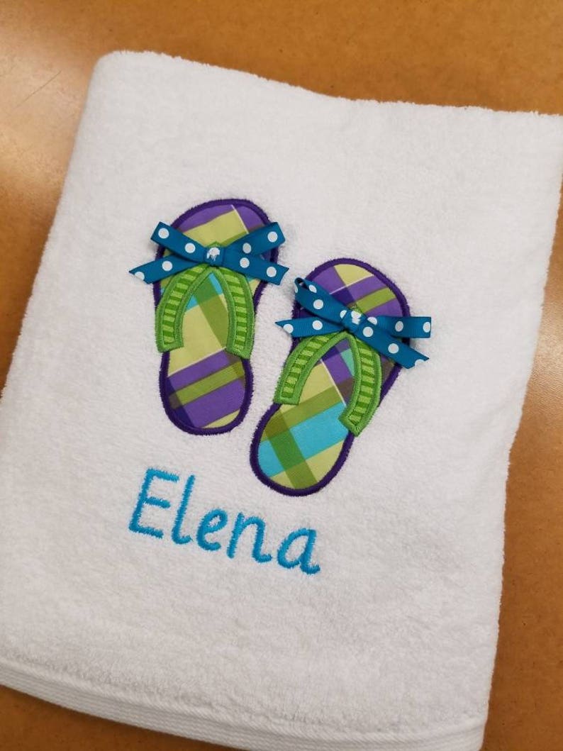May include: White bath towel with embroidered green and purple flip flops with blue bows and white polka dots. The name "Elena" is embroidered below the flip flops.