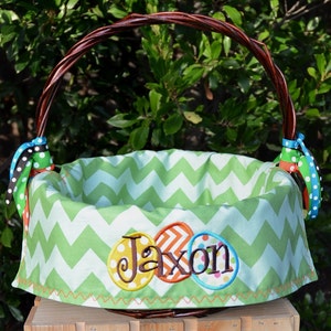 May include: A green and white chevron patterned Easter basket with a brown wicker handle. The basket has a personalized embroidered design with the name "Jaxon" in colorful letters.