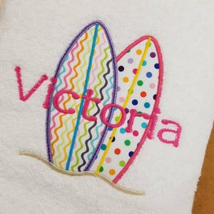 May include: White towel with two colorful surfboards embroidered on it. The surfboards have a pink outline and are decorated with colorful zigzags, dots, and stripes. The name "Victoria" is embroidered in pink letters on the towel.