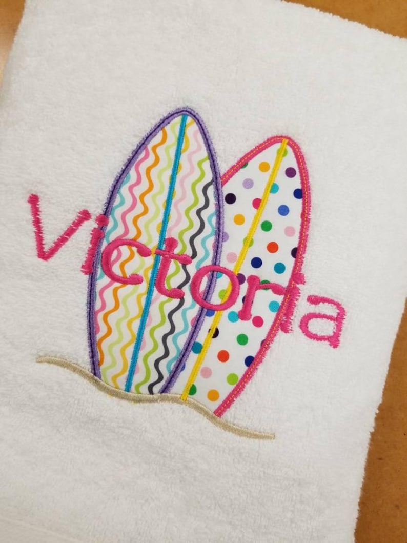 May include: White towel with two colorful surfboards embroidered on it. The surfboards have a pink outline and are decorated with colorful zigzags, dots, and stripes. The name "Victoria" is embroidered in pink letters on the towel.