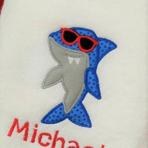 May include: White bath towel with a blue and gray shark wearing red sunglasses and the name Michael embroidered in red.