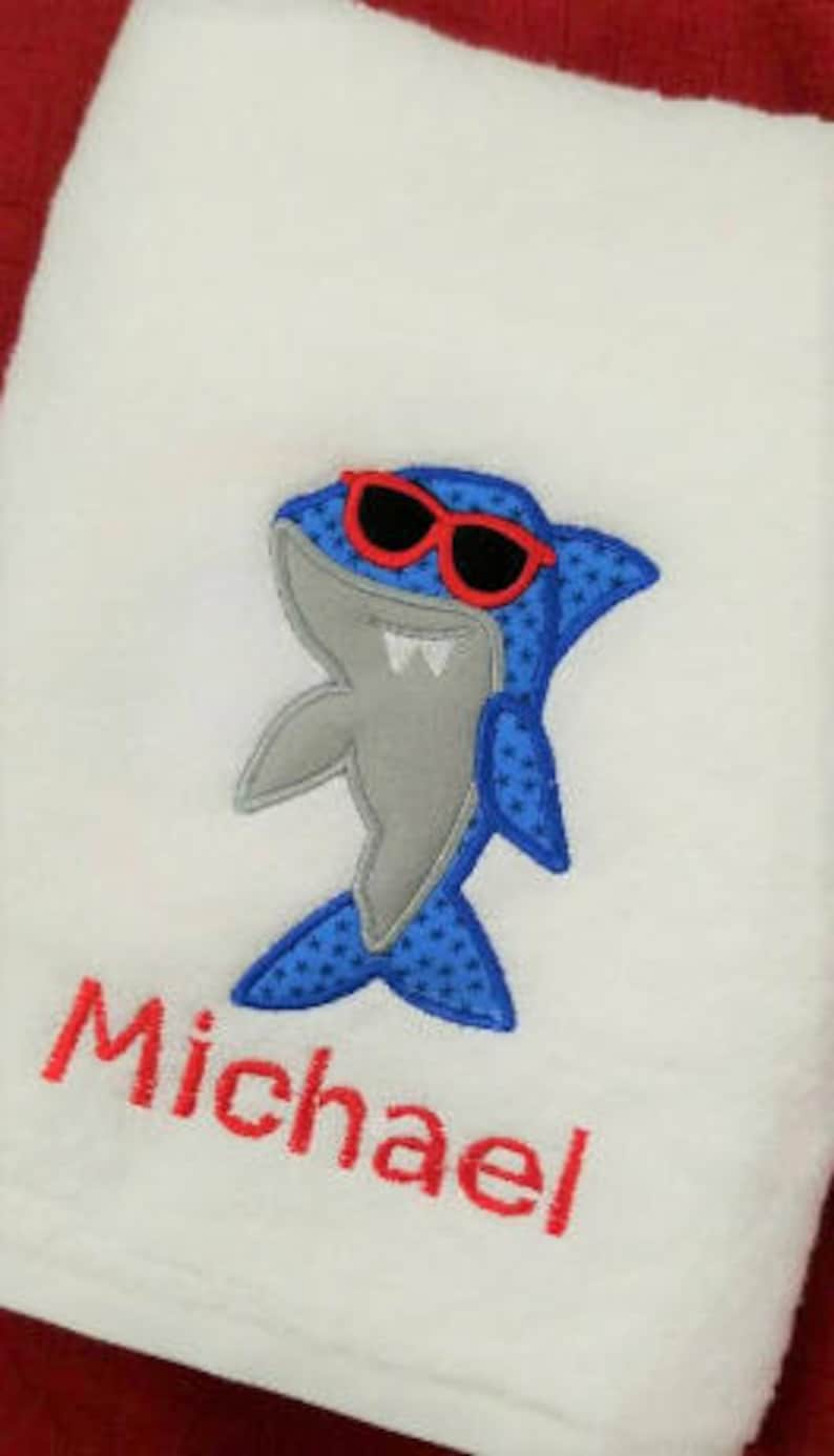 May include: White bath towel with a blue and gray shark wearing red sunglasses and the name Michael embroidered in red.