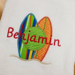 May include: White beach towel with a colorful embroidered design of two surfboards and a green circle with the name "Benjamin" in red.