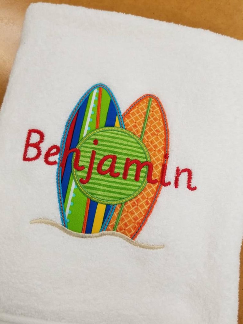 May include: White beach towel with a colorful embroidered design of two surfboards and a green circle with the name "Benjamin" in red.