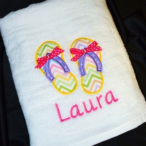 May include: White beach towel with embroidered flip flops and the name "Laura" in pink.