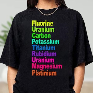 May include: A black short-sleeved t-shirt featuring a vertical list of chemical elements printed in vibrant colors. The text reads Fluorine, Uranium, Carbon, Potassium, Titanium, Rubidium, Uranium, Magnesium, and Platinium in a rainbow of hues.