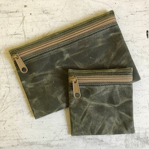May include: Two green canvas zippered pouches. The larger pouch is lying flat with the zipper facing up. The smaller pouch is standing upright with the zipper facing up. Both pouches have a brown zipper.