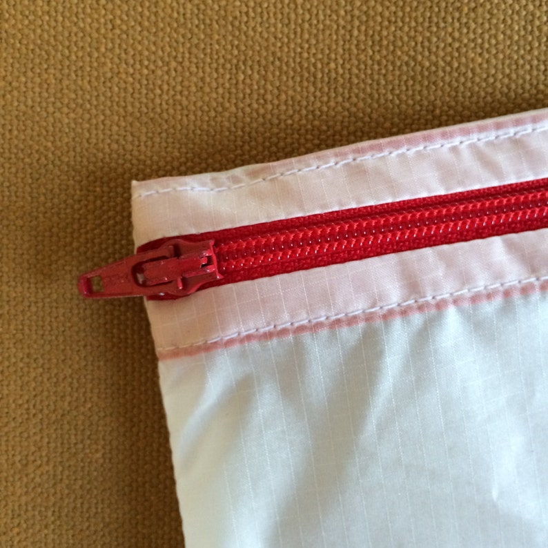 May include: A white zippered pouch with a red zipper. The pouch is made of a thin, lightweight material.