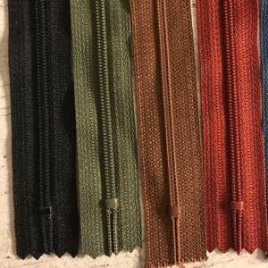 May include: Five zipper tapes in different colors: black, green, brown, red, and blue. The zippers are all closed and have a metal slider.