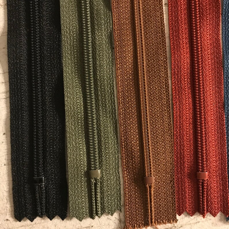 May include: Five zipper tapes in different colors: black, green, brown, red, and blue. The zippers are all closed and have a metal slider.