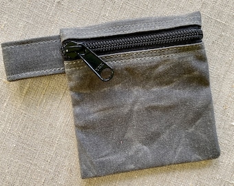 May include: A small gray zippered pouch with a black zipper pull. The pouch has a flap with a strap that can be used to attach it to something.