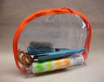 May include: A clear plastic travel bag with an orange zipper and trim. The bag contains a hairbrush, a hair clip, and a tube of lotion.