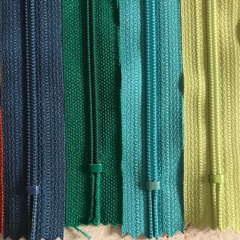 May include: Five zipper tapes in different colors: dark blue, green, teal, light blue, and light green. The zipper tapes are all the same size and have a smooth texture.