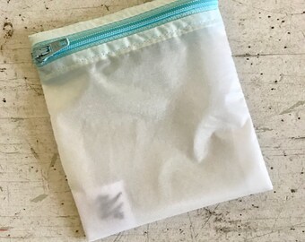 May include: A white mesh laundry bag with a light blue zipper. The bag is folded and lying on a light gray surface.
