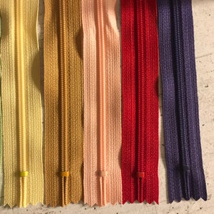 May include: A close-up of seven different colored zippers. The zippers are arranged in a row from left to right. The colors are: light green, yellow, gold, pink, red, purple, and dark purple.