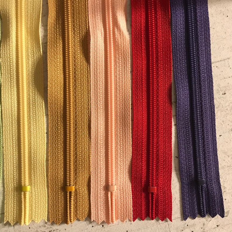 May include: A close-up of seven different colored zippers. The zippers are arranged in a row from left to right. The colors are: light green, yellow, gold, pink, red, purple, and dark purple.
