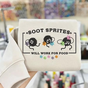 May include: A beige sweatshirt featuring the text "SOOT SPRITES. WILL WORK FOR FOOD" in a black rectangular frame. The design includes three cartoon soot sprites holding colorful stars. The sweatshirt's sleeve is visible.