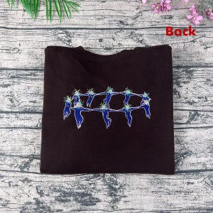May include: A dark brown sweatshirt featuring an embroidered design of figures holding hands in a circle. The figures are blue with white outlines and have colorful, spiky hair. The word "Back" is printed in red.