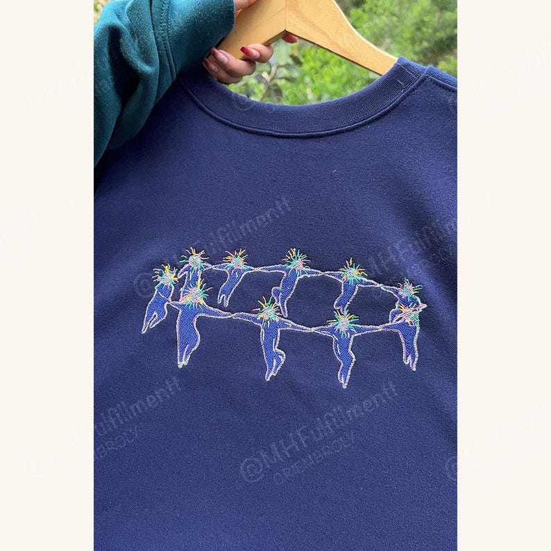 May include: Navy blue sweatshirt featuring an embroidered design of figures holding hands in a circle. The figures are blue with colorful accents. The sweatshirt is hanging on a wooden hanger.