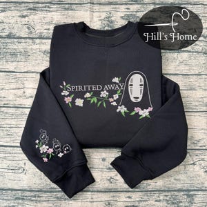 Może przedstawiać: Czarna bluza z haftowanym wzorem z filmu "Spirited Away". Wzór zawiera napis "Spirited Away", postać i kwiatowe akcenty w kolorze białym, różowym i zielonym. Rękaw ma również mały haftowany wzór.