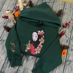 May include: Dark green hoodie featuring a colorful graphic of a character with a white mask and floral accents. The hoodie has a drawstring hood and embroidered details on the sleeve. The design is centered on the front of the sweatshirt.