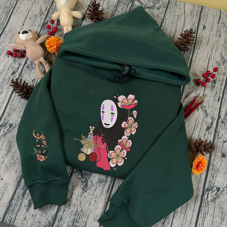 May include: Dark green hoodie featuring a colorful graphic of a character with a white mask and floral accents. The hoodie has a drawstring hood and embroidered details on the sleeve. The design is centered on the front of the sweatshirt.