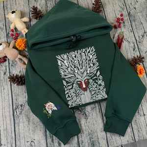 May include: A dark green hoodie with a hood and drawstring. The front features a white and red graphic of a face with a tree-like design. The left sleeve has an embroidered design. The hoodie is made of a soft material.