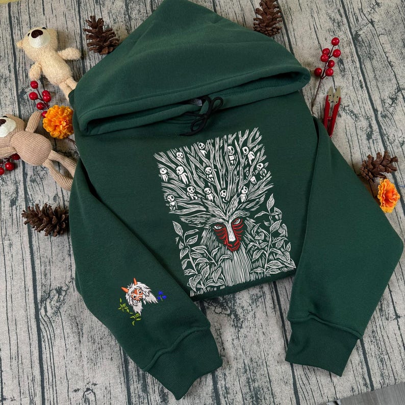 May include: A dark green hoodie with a hood and drawstring. The front features a white and red graphic of a face with a tree-like design. The left sleeve has an embroidered design. The hoodie is made of a soft material.