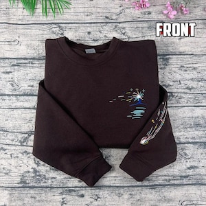 May include: A dark brown sweatshirt with embroidered celestial designs. The design includes a sun or star, with colorful lines and a shooting star on the sleeve. The word "FRONT" is printed in white.