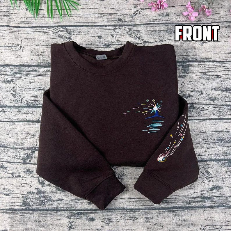 May include: A dark brown sweatshirt with embroidered celestial designs. The design includes a sun or star, with colorful lines and a shooting star on the sleeve. The word "FRONT" is printed in white.