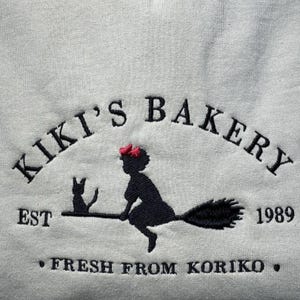 May include: White sweatshirt with an embroidered design. The design features the text "Kiki's Bakery" above a silhouette of a witch on a broom with a black cat. The text "EST 1989" and "FRESH FROM KORIKO" are also included.